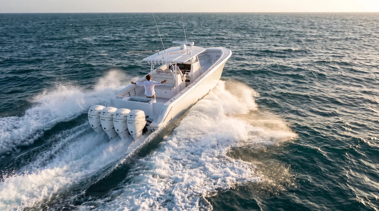 A white center-console boat with four outboard engines carving a wake across open ocean at sunset, a passenger standing at the stern with arms spread wide
